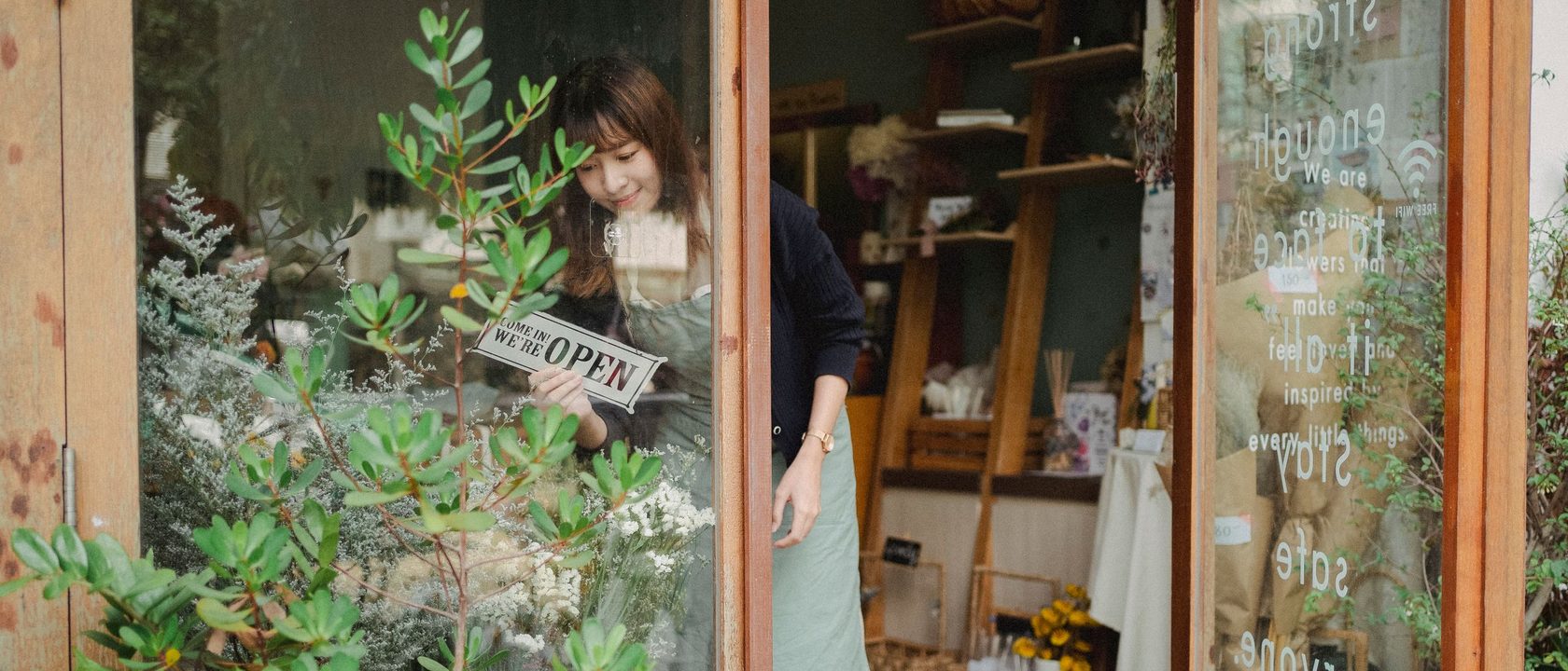 花店店員站在敞開的玻璃門邊，把「營業中」的牌子掛上門內，門口擺著盆栽。